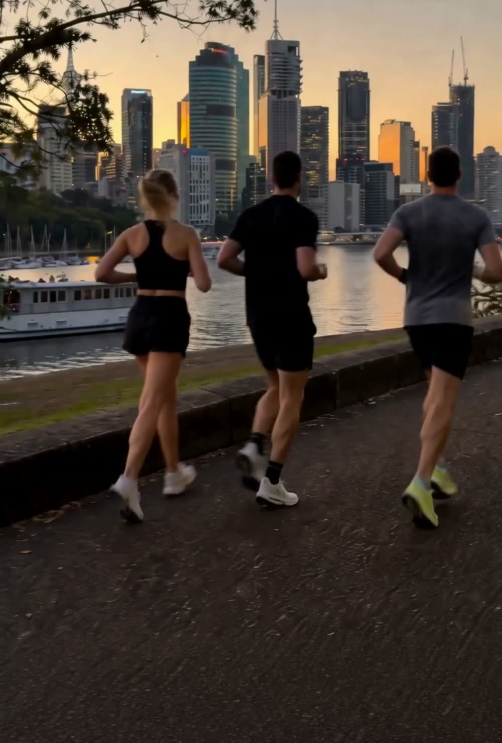 Three runners along the Brisbane River boardwalk at sunset, city skyline ahead